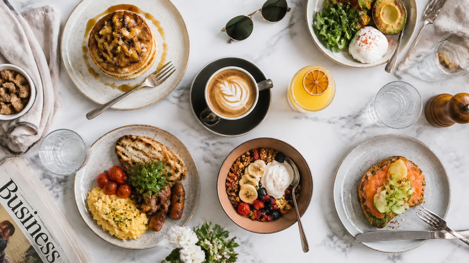 A top-down bird's-eye flat-lay of a vibrant gourmet brunch spread — pancakes, berries, coffee, avocado toast — neatly arranged on a marble table, bright natural light, editorial food photography, crisp.